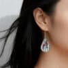 A close-up side profile of a woman with dark, shoulder-length hair wearing a teardrop-shaped dangle earring. The silver-framed pendant features a black and white scene of a birch forest with one or two vibrant red cardinals perched among the branches, giving it a winter or nature theme.
