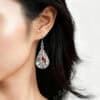 A close-up side profile of a woman with dark, shoulder-length hair wearing a teardrop-shaped dangle earring. The silver-framed pendant features a black and white scene of a birch forest with one or two vibrant red cardinals perched among the branches, giving it a winter or nature theme.