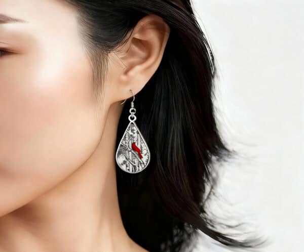 A close-up side profile of a woman with dark, shoulder-length hair wearing a teardrop-shaped dangle earring. The silver-framed pendant features a black and white scene of a birch forest with one or two vibrant red cardinals perched among the branches, giving it a winter or nature theme.