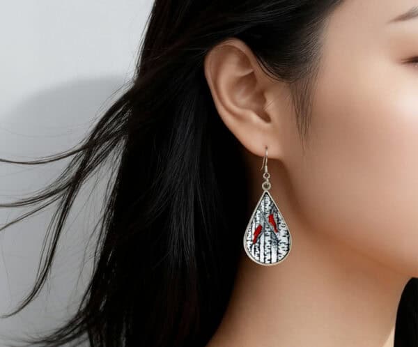 A close-up side profile of a woman with dark, shoulder-length hair wearing a teardrop-shaped dangle earring. The silver-framed pendant features a black and white scene of a birch forest with one or two vibrant red cardinals perched among the branches, giving it a winter or nature theme.