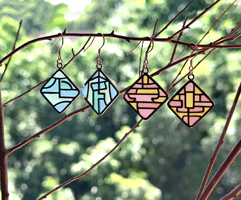 “Peace & Joy” plique-à-jour enamel earrings in soft blue and pink tones, featuring geometric stained-glass patterns, hanging on a branch with a blurred green nature background.The earrings feature geometric stained-glass-style designs framed in dark metal, showcasing delicate craftsmanship and vibrant translucent enamel.