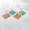 Two pairs of handcrafted plique-à-jour enamel earrings in square silver frames, featuring abstract line designs inspired by Chinese characters for “Peace” and “Joy,” with soft blue, pink, and golden hues glowing through openwork patterns, displayed on a white background with soft white feathers.