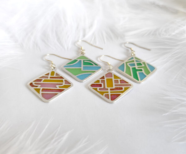 Two pairs of handcrafted plique-à-jour enamel earrings in square silver frames, featuring abstract line designs inspired by Chinese characters for “Peace” and “Joy,” with soft blue, pink, and golden hues glowing through openwork patterns, displayed on a white background with soft white feathers.