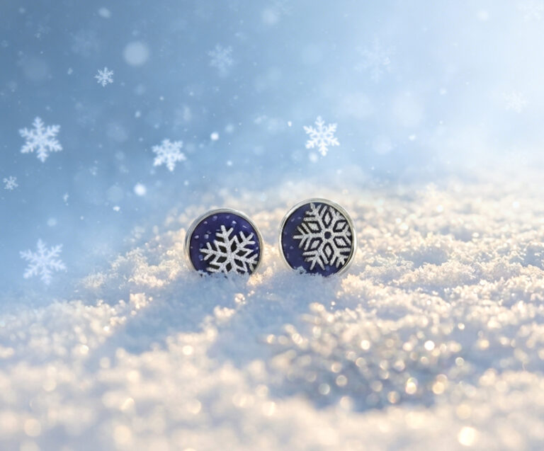 A pair of "Winter Sprite" stud earrings featuring silver frosted enamel snowflakes on a deep blue base, nestled in soft, sparkling white snow under warm, golden winter sunlight with a dreamy bokeh background.
