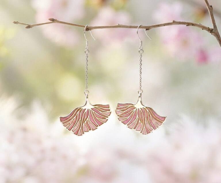 Pale lavender-pink poppy dangle earrings with silver edges hanging on a delicate branch, with soft-focus pink and white cherry blossoms in the background, creating a serene spring aesthetic.