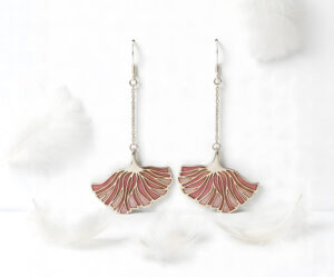 Poppy Grace Plique-à-jour enamel earrings in 925 sterling silver, featuring a delicate pink ginkgo-shaped fan design suspended on long silver chains, set against a blurry spring meadow background with soft sunlight.
