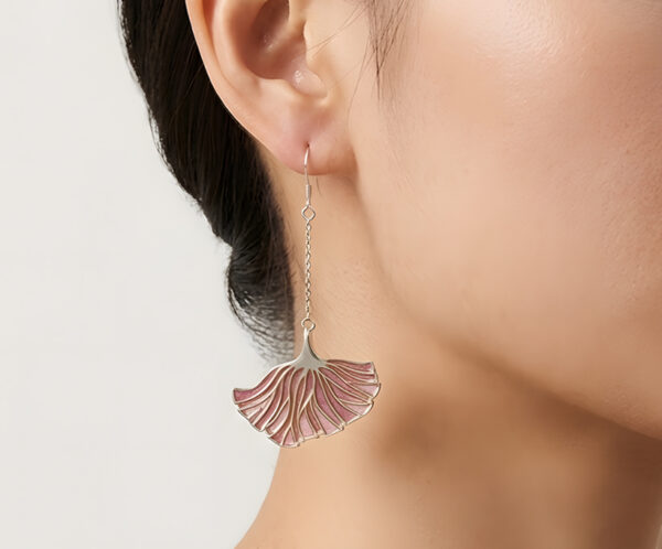 A close-up side profile of a model wearing the Poppy Grace Plique-à-jour enamel earrings. The light-as-wings pink enamel fans sway gracefully, catching the soft sunlight to reveal their semi-transparent texture against the model’s neck.